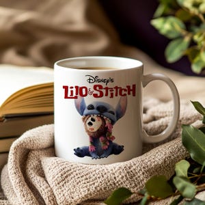 K&ouml;nnte beinhalten: Wei&szlig;e Keramik-Tasse mit Disney's Lilo & Stitch. Die Tasse zeigt Stitch, der eine Puppe h&auml;lt, mit dem Disney's Lilo & Stitch-Logo in Rot. Die Tasse hat einen gebogenen Griff und steht vor einem neutralen Hintergrund.