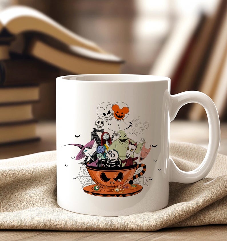May include: White ceramic mug featuring a Halloween-themed design. The design includes characters from The Nightmare Before Christmas, inside an orange teacup with a jack-o'-lantern face. The mug is set on a tan cloth.