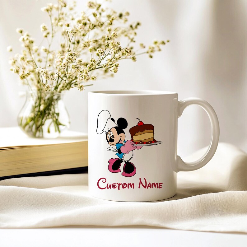 Pu&ograve; includere: Tazza in ceramica bianca con un personaggio dei cartoni animati che tiene in mano una torta. La tazza ha un manico e le parole "Custom Name" in corsivo rosso. Sullo sfondo un vaso di fiori bianchi. Capacit&agrave; di circa 300 ml.
