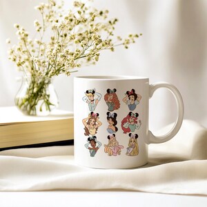 May include: White ceramic mug featuring cartoon illustrations of Disney princesses wearing Minnie Mouse ears. The princesses are depicted in various poses and outfits. A vase of white flowers and a book are in the background.