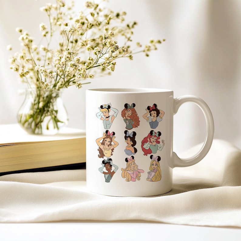 May include: White ceramic mug featuring cartoon illustrations of Disney princesses wearing Minnie Mouse ears. The princesses are depicted in various poses and outfits. A vase of white flowers and a book are in the background.