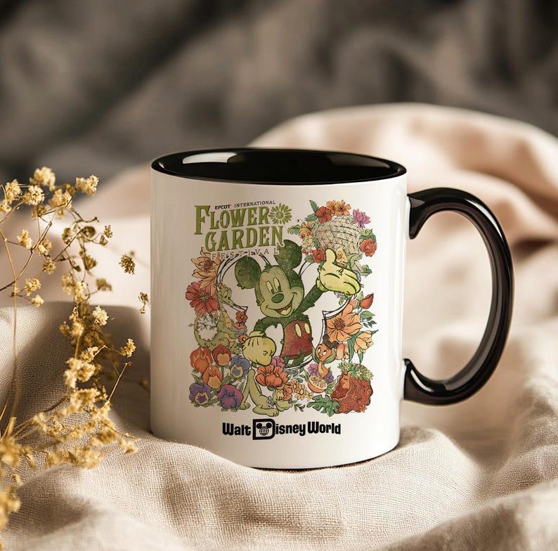 May include: White ceramic mug with a black interior and handle. The mug features a colourful graphic of Mickey Mouse surrounded by flowers, with the text "EPCOT INTERNATIONAL FLOWER GARDEN" and "Walt Disney World."