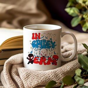 May include: White ceramic mug with the text "IN MY SPIDEY MOM ERA" in red and blue graffiti-style lettering. The mug features cartoon illustrations of Spider-Man characters. The mug is filled with a brown liquid.