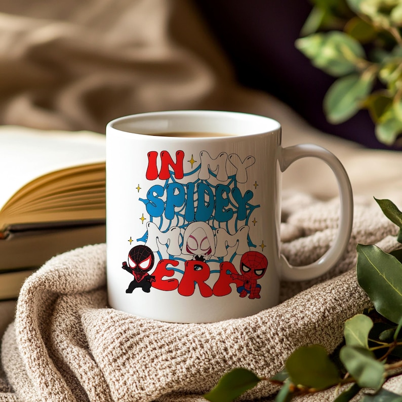 May include: White ceramic mug with the text "IN MY SPIDEY MOM ERA" in red and blue graffiti-style lettering. The mug features cartoon illustrations of Spider-Man characters. The mug is filled with a brown liquid.