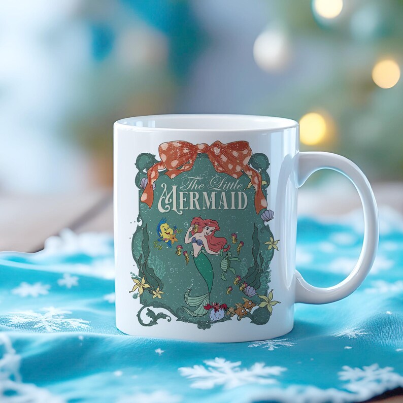 Pode incluir: Caneca de cer&acirc;mica branca com um design de "A Pequena Sereia". O design inclui Ariel, Linguado e Sebasti&atilde;o, com o t&iacute;tulo em uma moldura decorativa. Uma fita vermelha com bolinhas brancas est&aacute; no topo.