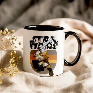 May include: White ceramic mug with a black interior and handle. The mug features a Star Wars logo and a cartoon character with a lightsaber. The design includes a retro colour scheme of orange, brown, and yellow.