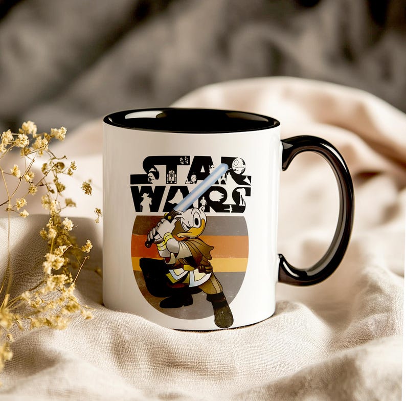 May include: White ceramic mug with a black interior and handle. The mug features a Star Wars logo and a cartoon character with a lightsaber. The design includes a retro colour scheme of orange, brown, and yellow.