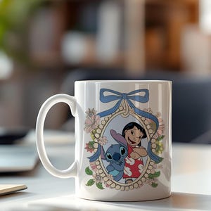 May include: White ceramic mug featuring a cartoon illustration of Lilo and Stitch. The design is framed by a floral wreath and a blue ribbon. The mug has a curved handle.