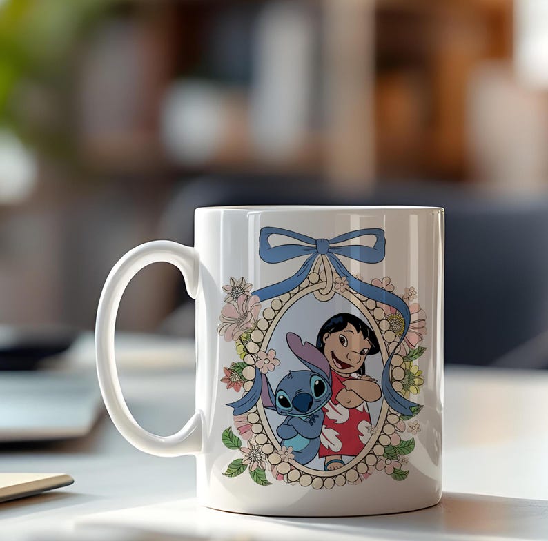 May include: White ceramic mug featuring a cartoon illustration of Lilo and Stitch. The design is framed by a floral wreath and a blue ribbon. The mug has a curved handle.