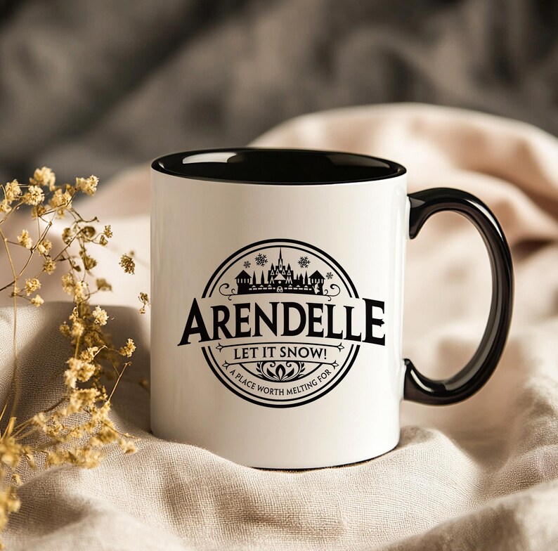 May include: White ceramic mug with a black interior and handle. The mug features a black circular design with the word "Arendelle" and the phrase "Let it snow! A place worth melting for."