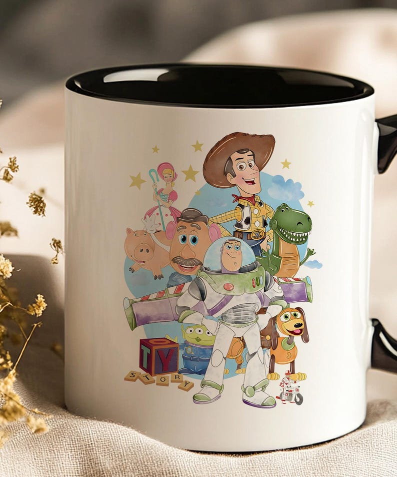 May include: White ceramic mug with a black interior and handle, featuring a colourful graphic of characters from the animated film Toy Story. The design includes Woody, Buzz Lightyear, and other beloved characters. The word "STORY" is also visible.
