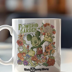 May include: White ceramic mug featuring Mickey Mouse surrounded by colourful flowers and the text "EPCOT INTERNATIONAL FLOWER GARDEN FESTIVAL" and "Walt Disney World". The design includes various floral elements and cartoon characters.