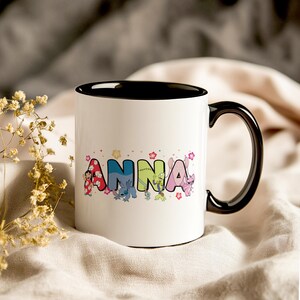 May include: White ceramic mug with a black interior and handle. The word "ANNA" is printed on the mug in colorful letters, each with a different character design. Floral accents and character illustrations surround the name.
