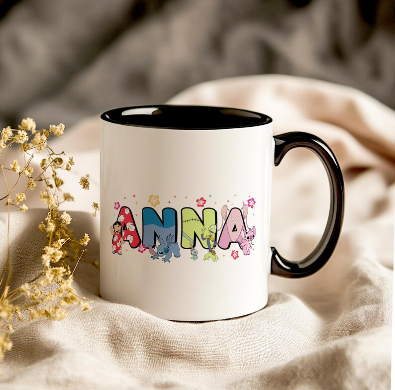 May include: White ceramic mug with a black interior and handle. The word "ANNA" is printed on the mug in colorful letters, each with a different character design. Floral accents and character illustrations surround the name.