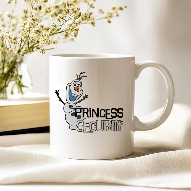 May include: White ceramic mug featuring a cartoon snowman character with the text "PRINCESS SECURITY" in black. The mug has a curved handle and is set against a soft, neutral background. A vase of white flowers and a book are visible.