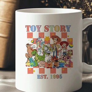 May include: White ceramic mug with a colorful graphic of the Toy Story characters, including Woody, Buzz Lightyear, Jessie, and Rex. The text "TOY STORY EST. 1995" is printed below the characters.