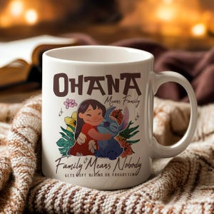 May include: White ceramic mug with the words "Ohana Means Family" in brown lettering. The mug features a cartoon illustration of a girl hugging a blue alien-like creature, surrounded by flowers. The phrase "Family Means Nobody Gets Left Behind or Forgotten" is also printed on the mug.