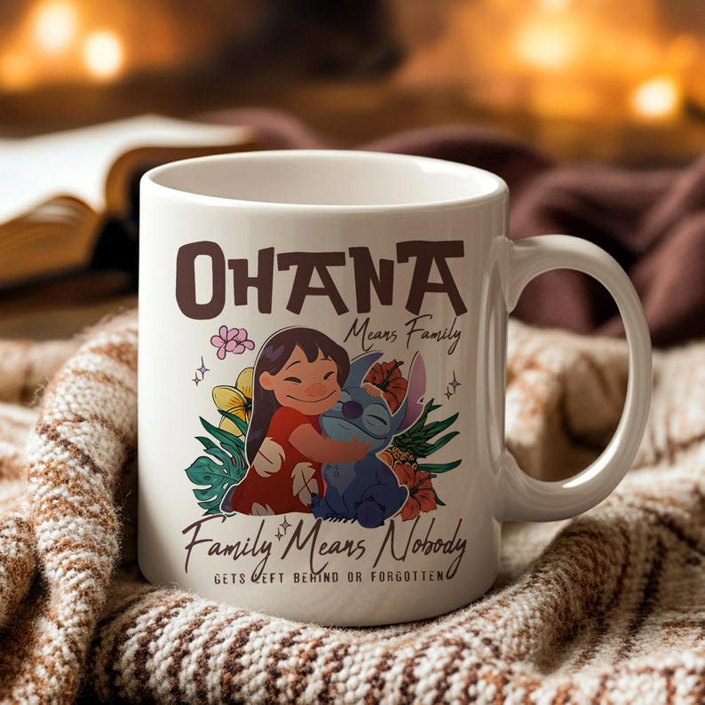 May include: White ceramic mug with the words "Ohana Means Family" in brown lettering. The mug features a cartoon illustration of a girl hugging a blue alien-like creature, surrounded by flowers. The phrase "Family Means Nobody Gets Left Behind or Forgotten" is also printed on the mug.