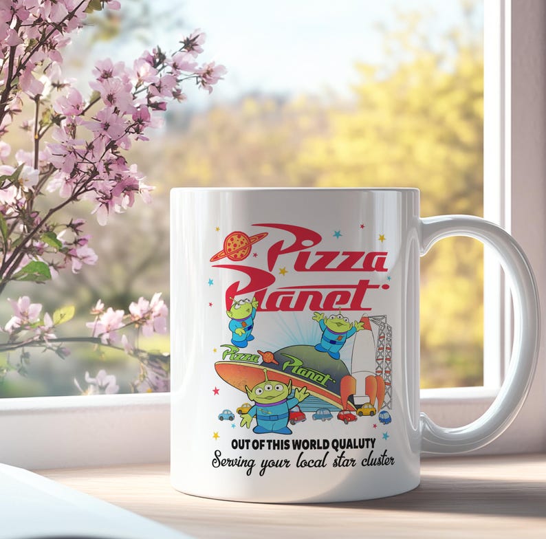 May include: White ceramic mug with a colorful image of the Pizza Planet restaurant from the Toy Story movie. The image features the restaurant's logo, a rocket ship, and three alien characters. The text on the mug reads "Pizza Planet", "Out of this world quality", and "Serving your local star cluster".