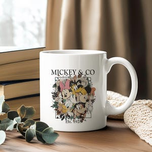 May include: White ceramic mug with a colorful illustration of Mickey Mouse, Minnie Mouse, Donald Duck, and Goofy surrounded by flowers. The text "Mickey & Co. Est. 1928" is printed on the mug.