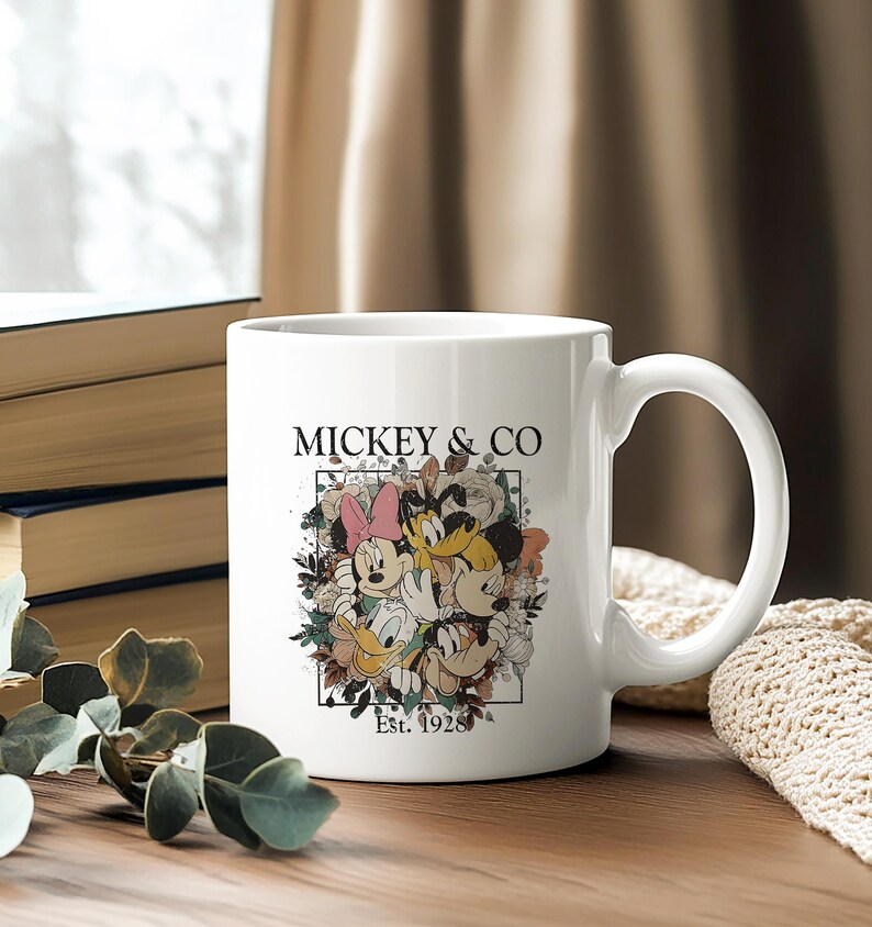 May include: White ceramic mug with a colorful illustration of Mickey Mouse, Minnie Mouse, Donald Duck, and Goofy surrounded by flowers. The text "Mickey & Co. Est. 1928" is printed on the mug.