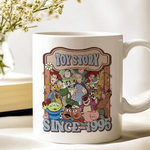 K&ouml;nnte beinhalten: Wei&szlig;e Keramik-Tasse mit einem Retro-Grafikdesign von Toy Story-Charakteren. Das Design enth&auml;lt den Text "TOY STORY" und "SINCE 1995". Die Tasse hat einen gebogenen Griff und ist f&uuml;r hei&szlig;e oder kalte Getr&auml;nke geeignet.