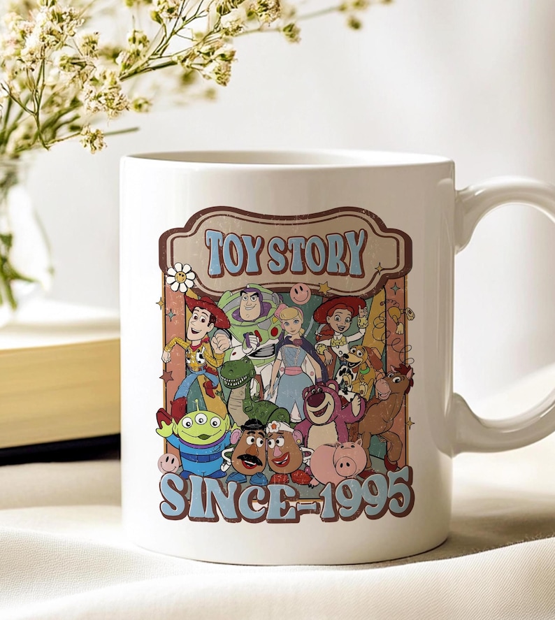 K&ouml;nnte beinhalten: Wei&szlig;e Keramik-Tasse mit einem Retro-Grafikdesign von Toy Story-Charakteren. Das Design enth&auml;lt den Text "TOY STORY" und "SINCE 1995". Die Tasse hat einen gebogenen Griff und ist f&uuml;r hei&szlig;e oder kalte Getr&auml;nke geeignet.