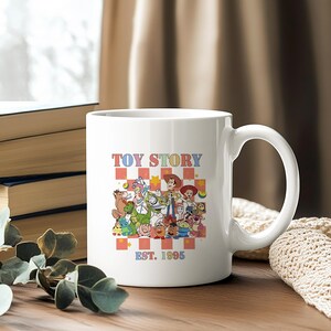 May include: White ceramic mug with a colorful illustration of characters from the Toy Story franchise. The mug has the text "TOY STORY" and "EST. 1995" printed on it.