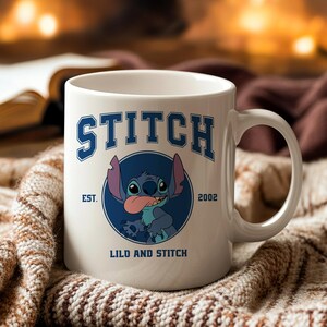 Puede incluir: Taza de cer&aacute;mica blanca con la palabra "STITCH" en letras azules y un gr&aacute;fico del personaje Stitch. La taza tiene el texto "EST. 2002" y "LILO AND STITCH". La taza tiene un asa curva y est&aacute; sobre un fondo c&aacute;lido y borroso.