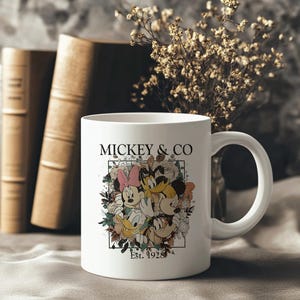 May include: White ceramic mug with a colorful illustration of Mickey Mouse, Minnie Mouse, Donald Duck, and Goofy surrounded by flowers. The text "MICKEY & CO. Est. 1928" is printed on the mug.