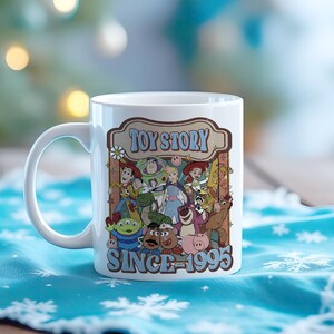 K&ouml;nnte beinhalten: Wei&szlig;e Keramik-Tasse mit einer farbenfrohen Grafik von Toy Story-Charakteren. Das Design enth&auml;lt den Text "TOY STORY" und "SINCE 1995". Die Tasse hat einen gebogenen Griff und steht vor einem verschwommenen Hintergrund.