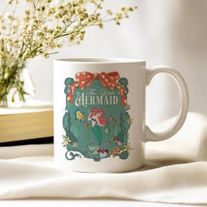 Pode incluir: Caneca de cer&acirc;mica branca com um design de "A Pequena Sereia". O design inclui uma sereia ruiva, um la&ccedil;o e as palavras "The Little Mermaid". A caneca tem uma al&ccedil;a curva e est&aacute; sobre um fundo macio e neutro.