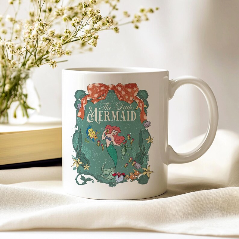 Pode incluir: Caneca de cer&acirc;mica branca com um design de "A Pequena Sereia". O design inclui uma sereia ruiva, um la&ccedil;o e as palavras "The Little Mermaid". A caneca tem uma al&ccedil;a curva e est&aacute; sobre um fundo macio e neutro.