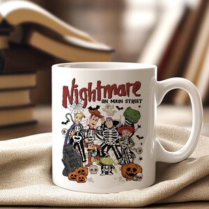 May include: White ceramic mug featuring a Halloween-themed design with the text "Nightmare on Main Street." The design includes cartoon characters dressed as skeletons, pumpkins, and a tombstone. The mug has a curved handle.