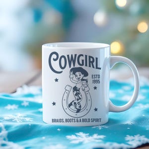 Pu&ograve; includere: Tazza in ceramica bianca con grafica nera di una cowgirl all'interno di un ferro di cavallo, la parola "COWGIRL" sopra, e il testo "ESTD 1995" e "BRAIDS, BOOTS & A BOLD SPIRIT" sotto. La tazza ha un manico bianco ed &egrave; su una superficie blu.
