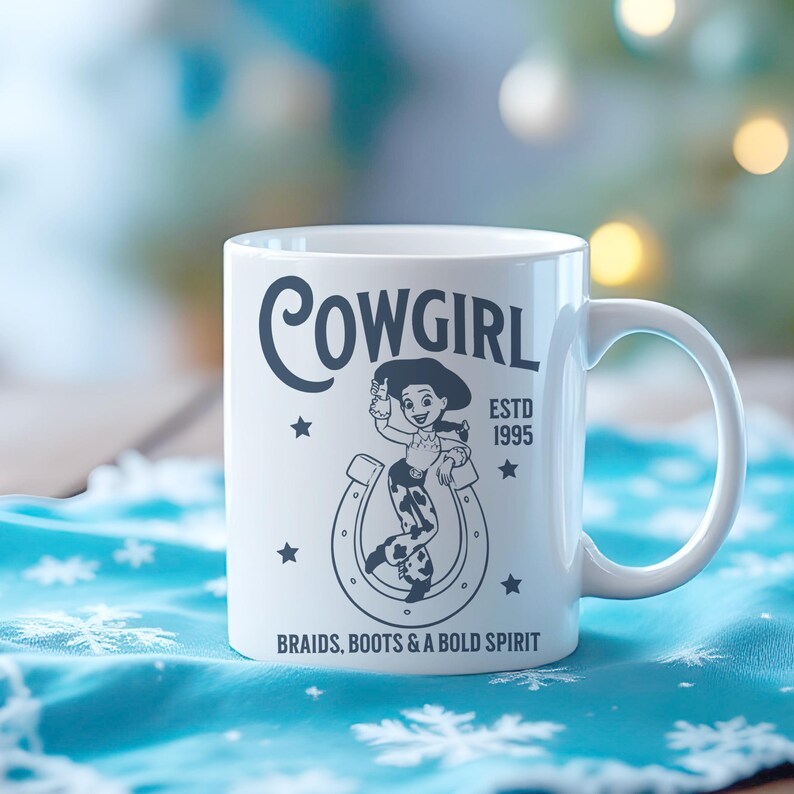 Pu&ograve; includere: Tazza in ceramica bianca con grafica nera di una cowgirl all'interno di un ferro di cavallo, la parola "COWGIRL" sopra, e il testo "ESTD 1995" e "BRAIDS, BOOTS & A BOLD SPIRIT" sotto. La tazza ha un manico bianco ed &egrave; su una superficie blu.