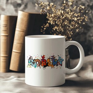 May include: White ceramic mug with the name "ROBERT" in colorful, cartoon-style letters. The letters are decorated with various cartoon characters. The mug has a curved handle and is set against a blurred background with books and flowers.