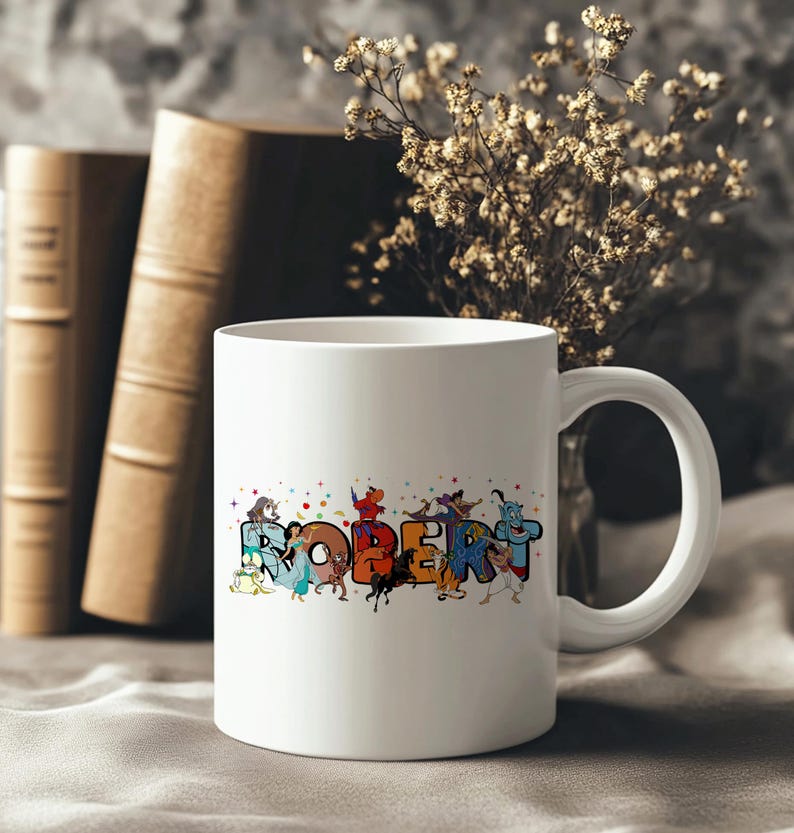 May include: White ceramic mug with the name "ROBERT" in colorful, cartoon-style letters. The letters are decorated with various cartoon characters. The mug has a curved handle and is set against a blurred background with books and flowers.