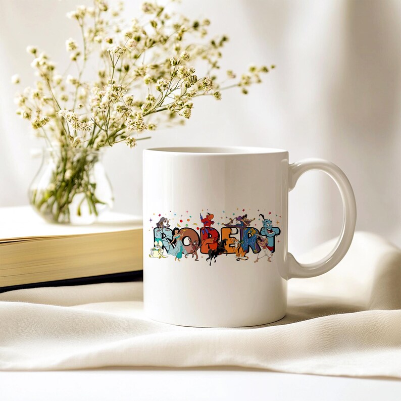 May include: White ceramic mug with the name "ROBERT" in colorful, cartoon-style lettering. The letters are adorned with various animated characters. A small vase of white flowers and a book are in the background.