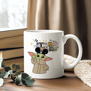 Może przedstawiać: Biały ceramiczny kubek z kresk&oacute;wkowym wizerunkiem Grogu, znanego r&oacute;wnież jako Baby Yoda, w uszach Myszki Miki. Kubek ma logo Gwiezdnych Wojen i inne grafiki związane z Gwiezdnymi Wojnami. Kubek ma duży uchwyt.