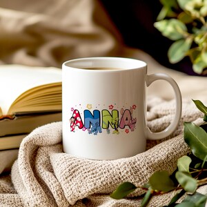 May include: White ceramic mug with the name "Anna" in colorful, cartoon-style letters. The letters are decorated with floral accents and character illustrations. The mug is filled with a dark liquid and rests on a beige knitted blanket.
