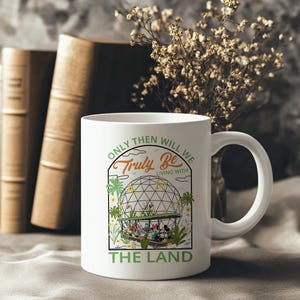 May include: White ceramic mug with a curved handle. The mug features a graphic of a geodesic dome with cartoon characters and the text "ONLY THEN WILL WE TRULY BE LIVING WITH THE LAND" in green and black. The mug is set against a blurred background.