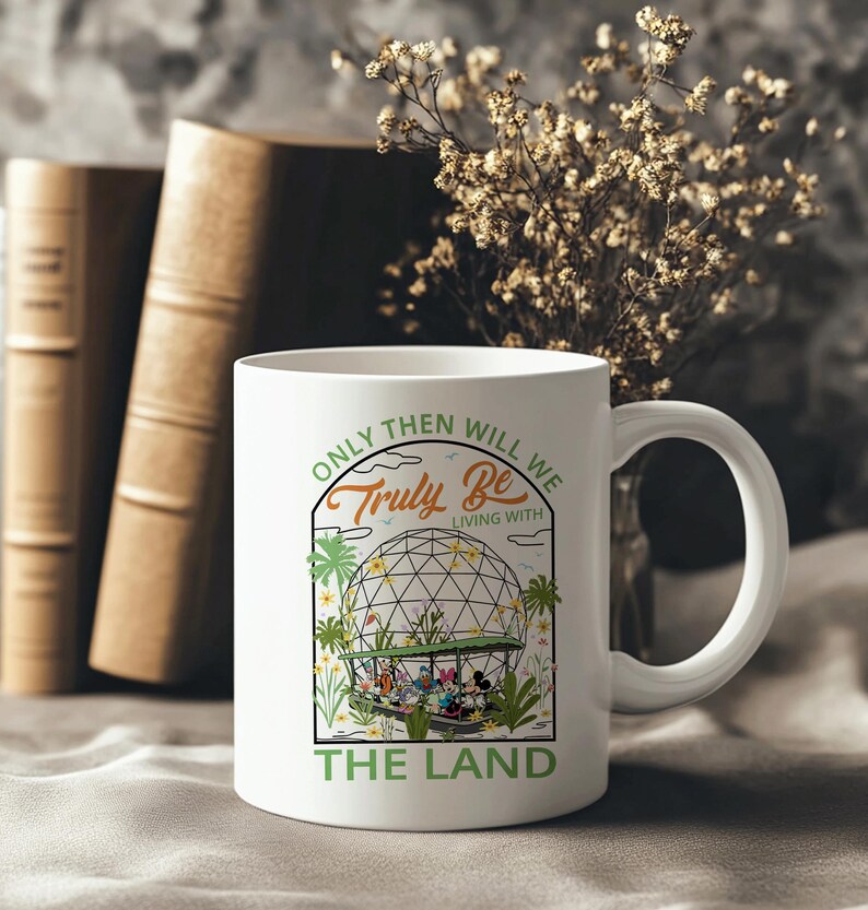 May include: White ceramic mug with a curved handle. The mug features a graphic of a geodesic dome with cartoon characters and the text "ONLY THEN WILL WE TRULY BE LIVING WITH THE LAND" in green and black. The mug is set against a blurred background.