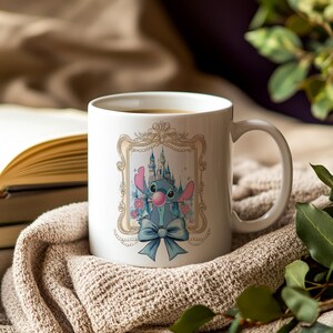 May include: White ceramic mug featuring a cartoon character with a castle backdrop. The character is blue with large ears and a pink bubble. A blue bow is at the bottom of the image. The mug is sitting on a beige knitted blanket.