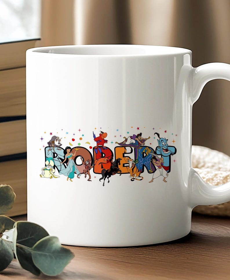 May include: White ceramic mug featuring the name "ROBERT" in colorful, cartoon-style letters. Each letter is decorated with characters from a popular animated movie. The mug has a curved handle and is set against a blurred background.