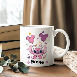 Pu&ograve; includere: Tazza in ceramica bianca con un personaggio dei cartoni animati rosa che tiene in mano palloncini rosa e viola a forma di Topolino. La tazza ha la scritta "Name" in nero, con stelle. Sfondo sfocato.