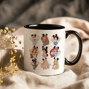 May include: White ceramic mug with a black interior and handle. The mug features cartoon illustrations of various princesses wearing mouse ears. The princesses are in different poses and have different hair colors. The mug is on a neutral background.