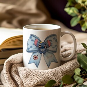 Pu&ograve; includere: Tazza in ceramica bianca con un fiocco blu a pois e un personaggio dei cartoni animati. Il personaggio ha una corona. Un fiore di ibisco rosa e la parola "Ohana" sono anche sul fiocco.