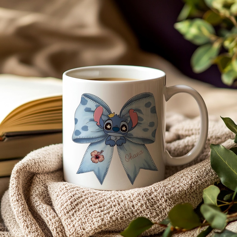 Pu&ograve; includere: Tazza in ceramica bianca con un fiocco blu a pois e un personaggio dei cartoni animati. Il personaggio ha una corona. Un fiore di ibisco rosa e la parola "Ohana" sono anche sul fiocco.