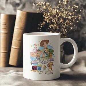 May include: White ceramic mug with a colourful illustration of characters from the animated film Toy Story. The design includes Woody, Buzz Lightyear, Rex, and other characters. The mug has a curved handle and is set against a neutral background.